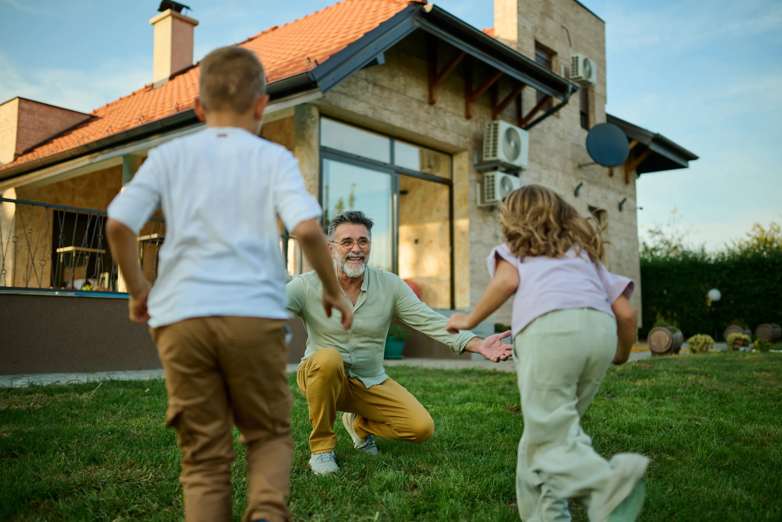 Grandfather kneeling on the lawn as two grandchildren run toward him